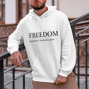 May include: White hoodie with a front pocket and a hood. The word "FREEDOM" is printed in black, with "inspired by American spirit" underneath. The hoodie is a casual garment.