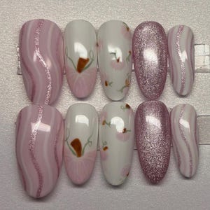 May include: A set of ten artificial nails in various designs. The nails feature a combination of pink glitter, white and pink swirl patterns, and floral designs with brown and pink accents. The nails are oval-shaped.
