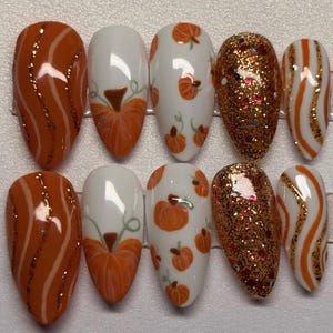 May include: A set of ten artificial nails with a fall theme. Designs include orange and gold glitter, orange and white stripes, and white nails with orange pumpkin illustrations. The nails are oval-shaped.
