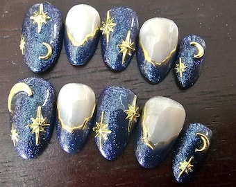 May include: Set of ten artificial nails with a celestial theme. The nails feature a dark blue glitter base with gold star and crescent moon accents. Some nails have a white center with gold trim.