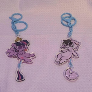May include: Two acrylic keychains with light blue clasps. The left keychain features a purple pony with a crown and a small tail charm. The right keychain features a light purple creature with dark patches and a small crescent moon charm.