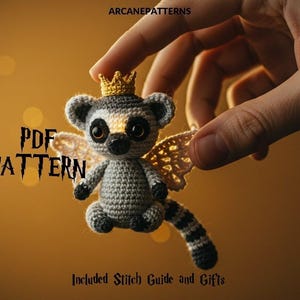May include: A crocheted lemur, adorned with a gold crown and wings, is held. The lemur features grey, white, and black colours, with large eyes. The text "PDF PATTERN" is visible. The background is a warm, blurred golden hue.