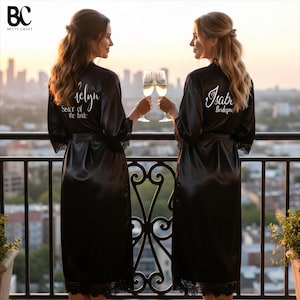 Könnte beinhalten: Zwei schwarze Satin-Morgenmäntel mit Spitzenbesatz an Ärmeln und Saum. Der linke Morgenmantel trägt den weißen Schriftzug 'Katelyn' und 'Sister of the bride', der rechte 'Isabel' und 'Bridesmaid'. Beide haben einen Bindegürtel.