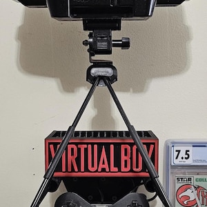 May include: A red and black Virtual Boy gaming console mounted on a black tripod stand. The device features the text VIRTUAL BOY on the front of the visor and the base unit. A black game controller with red buttons is attached to the front of the stand.