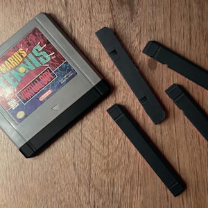 Könnte beinhalten: Eine graue Nintendo Virtual Boy-Spielkassette mit der Aufschrift "Mario's Tennis" und schwarzen Kunststoffkomponenten. Vier schwarze Kunststoffteile sind auf einer Holzoberfläche um die Kassette herum verstreut. Die Spielkassette hat ein rotes, blaues und weißes Etikett.
