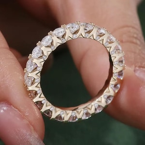 Peut inclure: Une bague d'éternité dorée ornée de nombreux diamants ronds et transparents, sertis côte à côte. La bague est tenue par une main, mettant en valeur son éclat et son design. Un bijou élégant.