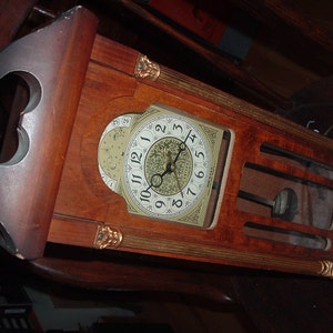 May include: A vintage wooden grandfather clock with a light-colored clock face featuring black numerals and ornate hands. The clock has a dark brown finish with decorative gold accents and a pendulum visible through a glass panel.