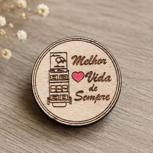 Pode incluir: Um broche circular de madeira com um gráfico gravado a laser de um carrinho de vários níveis e o texto "Melhor Vida de Sempre" ao lado de um pequeno coração rosa vibrante. O design é emoldurado por duas linhas circulares concêntricas.