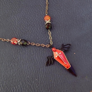 May include: A gothic-style necklace featuring a coffin pendant with a cross design. The pendant is red and black with small black wings. The necklace has a gold-colored chain with red, orange, and black beads.