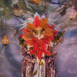May include: A whimsical, handcrafted figure with a face adorned with large, expressive eyes and a pointed nose. The figure's head is framed by vibrant red and orange leaf-like elements, with a beard of similar leaves. The figure is dressed in a patterned fabric.