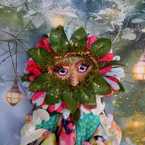 May include: A whimsical, handcrafted mask featuring a face with large, expressive eyes and a leafy beard. The mask is adorned with red, pink, and white petals, green leaves, and gold glitter accents. The mask is part of a costume.