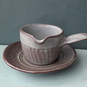 May include: A white ceramic gravy boat with a brown handle and a matching brown saucer. The boat has a textured surface and a spout for pouring.