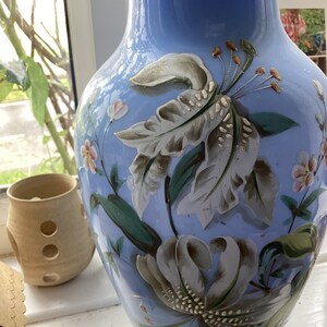May include: A light blue vase with hand-painted floral designs in white, gray, and green. The vase features a wide base and a slender neck. A small, tan ceramic container with circular cutouts is in the background.