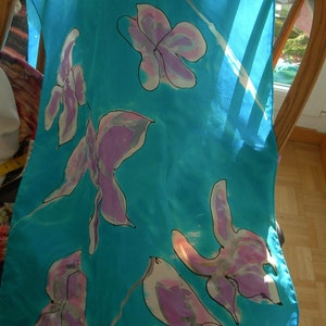 May include: A turquoise silk scarf with a floral pattern. The scarf features large, stylized flowers in shades of pink and purple, outlined in black. The scarf is draped over a wooden chair.