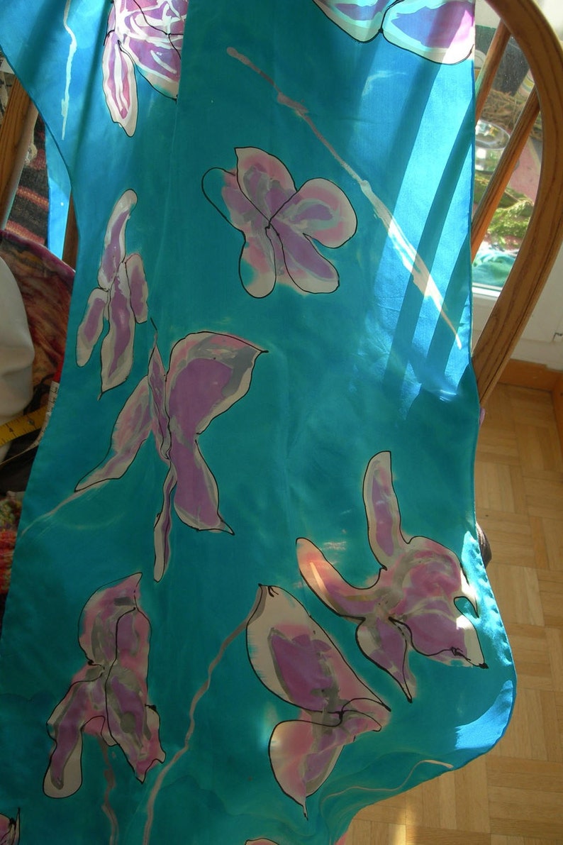 May include: A turquoise silk scarf with a floral pattern. The scarf features large, stylized flowers in shades of pink and purple, outlined in black. The scarf is draped over a wooden chair.