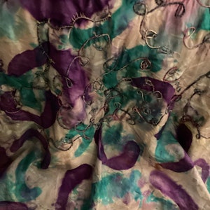 May include: Abstract textile with a watercolor effect. The fabric features a cream background with splashes of purple, teal, and pink. Silver thread embroidery adds a delicate, swirling pattern across the surface.