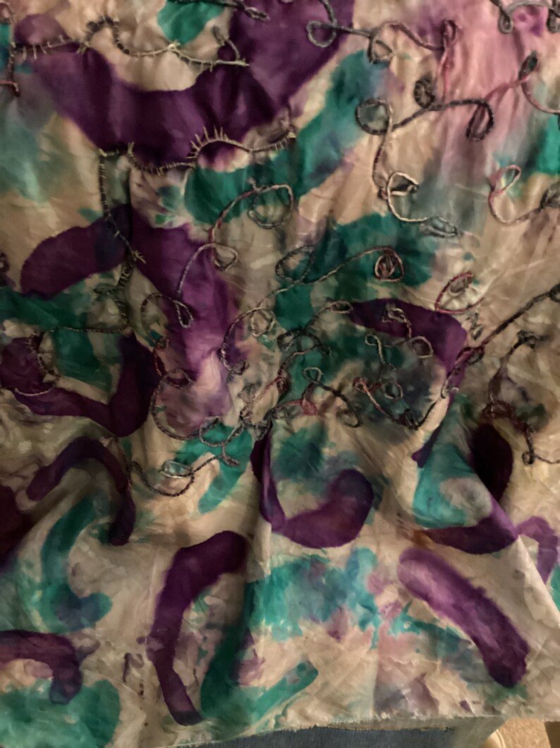 May include: Abstract textile with a watercolor effect. The fabric features a cream background with splashes of purple, teal, and pink. Silver thread embroidery adds a delicate, swirling pattern across the surface.