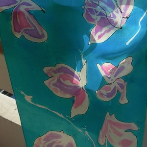 May include: A turquoise scarf with a floral pattern. The flowers are white with pink and purple accents and black outlines. The scarf is made of a lightweight fabric and is hanging in front of a natural background.