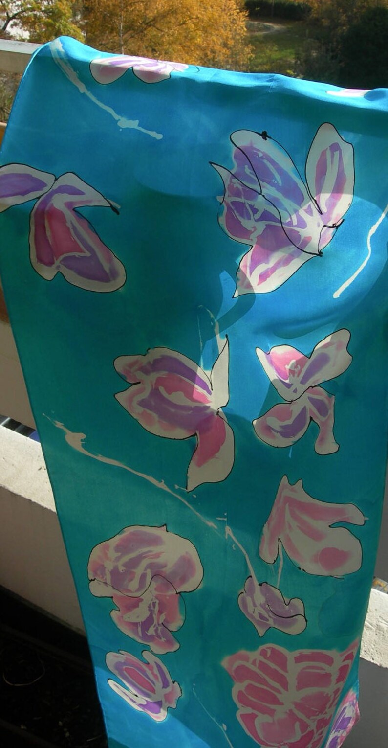 May include: A turquoise scarf with a floral pattern. The flowers are white with pink and purple accents and black outlines. The scarf is made of a lightweight fabric and is hanging in front of a natural background.