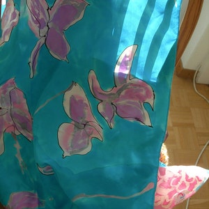 May include: A turquoise silk scarf with a floral pattern. The design features large, stylized flowers in shades of pink and purple with black outlines. The scarf is draped over a chair, with sunlight shining through.