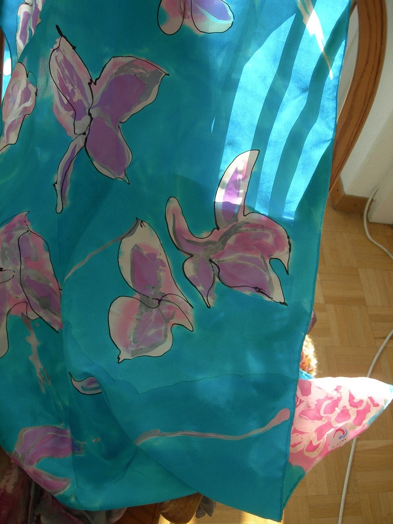 May include: A turquoise silk scarf with a floral pattern. The design features large, stylized flowers in shades of pink and purple with black outlines. The scarf is draped over a chair, with sunlight shining through.