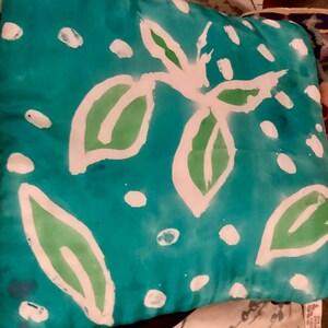 May include: Square decorative pillow with a teal background and a white and green leaf design. The leaves are outlined in white and filled with green. White polka dots are scattered across the pillow.