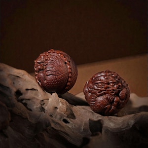 Op de afbeelding: Twee ingewikkeld gesneden, bolvormige houten sculpturen. De sculpturen hebben een dieprood-bruine kleur en hebben gedetailleerde drakenontwerpen. Ze rusten op een stuk verweerd hout tegen een donkerbruine achtergrond.