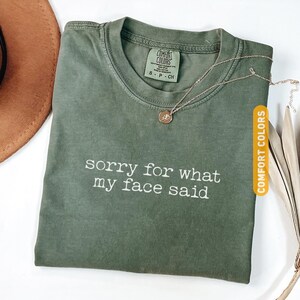 Pu&ograve; includere: Una maglietta verde muschio con la scritta bianca "sorry for what my face said". La maglietta ha un girocollo e maniche corte. Una collana d'oro con un piccolo ciondolo &egrave; appoggiata sulla maglietta. La maglietta &egrave; etichettata "Comfort Colors".
