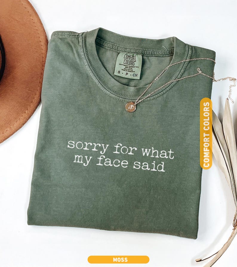 Pu&ograve; includere: Una maglietta verde muschio con la scritta bianca "sorry for what my face said". La maglietta ha un girocollo e maniche corte. Una collana d'oro con un piccolo ciondolo &egrave; appoggiata sulla maglietta. La maglietta &egrave; etichettata "Comfort Colors".
