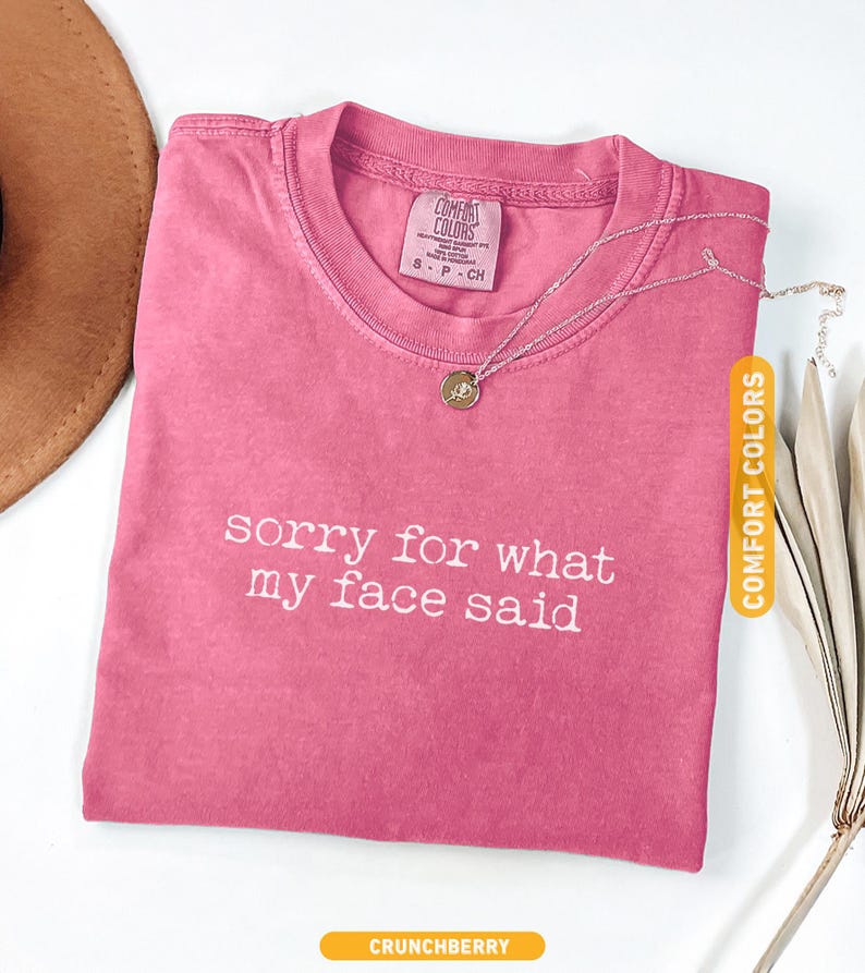 Pu&ograve; includere: Una maglietta rosa con la scritta bianca "sorry for what my face said". La maglietta ha uno scollo tondo e una piccola etichetta. Una collana d'argento con un ciondolo &egrave; sulla maglietta. La parola "CRUNCHBERRY" &egrave; in basso.