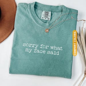 Pu&ograve; includere: Una maglietta verde chiaro con la scritta bianca "sorry for what my face said". Una collana color oro con un ciondolo poggia sulla maglietta. La maglietta &egrave; etichettata "Comfort Colors".
