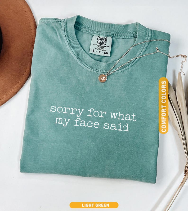 Pu&ograve; includere: Una maglietta verde chiaro con la scritta bianca "sorry for what my face said". Una collana color oro con un ciondolo poggia sulla maglietta. La maglietta &egrave; etichettata "Comfort Colors".