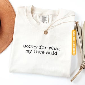Pu&ograve; includere: T-shirt color avorio con la scritta nera "sorry for what my face said". Una collana d'oro con un ciondolo &egrave; sulla maglietta. La maglietta &egrave; etichettata "Comfort Colors". Un cappello marrone &egrave; visibile nell'angolo in alto a sinistra.