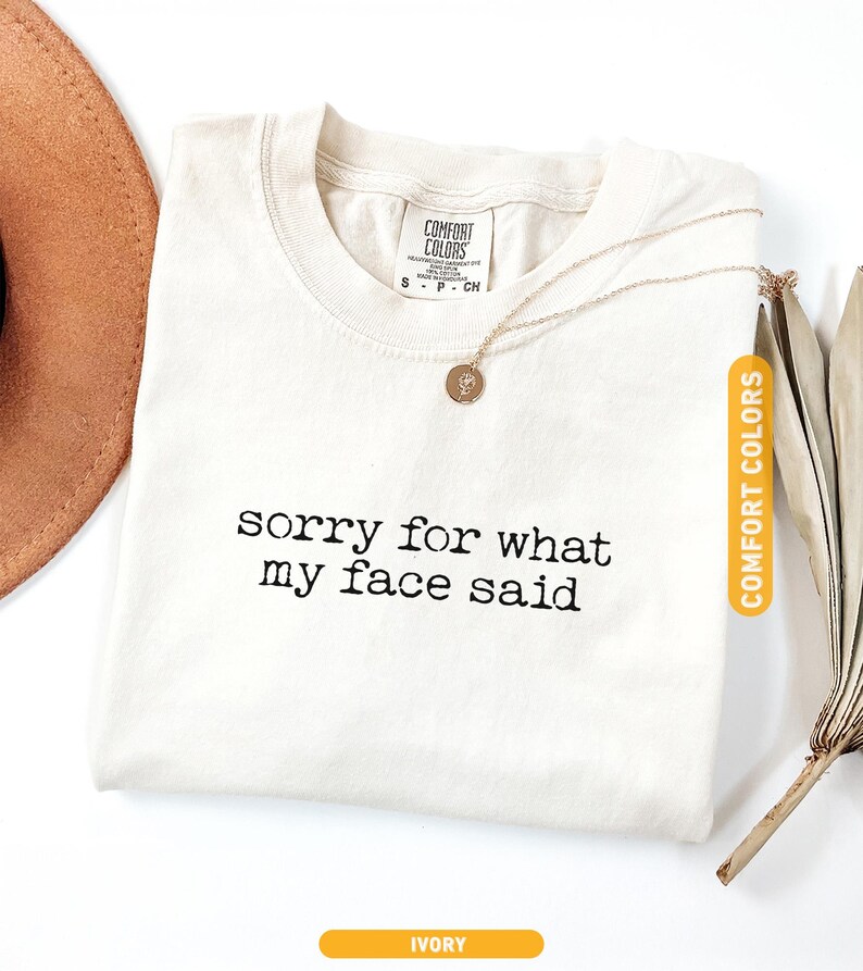 Pu&ograve; includere: T-shirt color avorio con la scritta nera "sorry for what my face said". Una collana d'oro con un ciondolo &egrave; sulla maglietta. La maglietta &egrave; etichettata "Comfort Colors". Un cappello marrone &egrave; visibile nell'angolo in alto a sinistra.