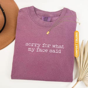 Pu&ograve; includere: T-shirt color bacca con la scritta bianca "sorry for what my face said". Una collana d'oro con un ciondolo rettangolare &egrave; appoggiata sulla maglietta. La maglietta &egrave; etichettata "Comfort Colors".