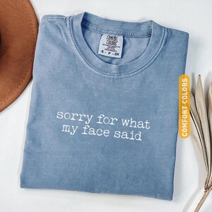 Pu&ograve; includere: T-shirt blu jeans con la scritta bianca "sorry for what my face said". La maglietta ha uno scollo tondo e una piccola etichetta con la scritta "COMFORT COLORS". Un cappello marrone e piante secche sullo sfondo.