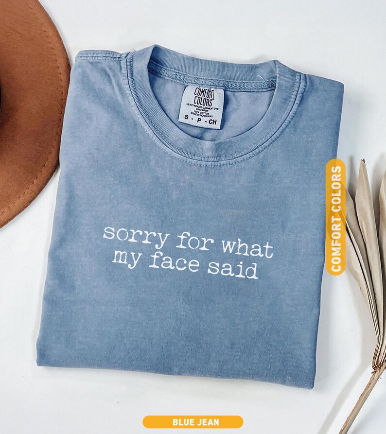 Pu&ograve; includere: T-shirt blu jeans con la scritta bianca "sorry for what my face said". La maglietta ha uno scollo tondo e una piccola etichetta con la scritta "COMFORT COLORS". Un cappello marrone e piante secche sullo sfondo.