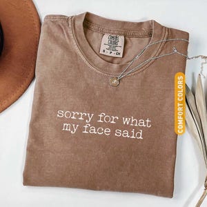 Pu&ograve; includere: T-shirt Comfort Colors color espresso con la scritta bianca "sorry for what my face said". Una collana d'argento con un ciondolo &egrave; appoggiata sulla maglietta. Un cappello marrone &egrave; visibile sullo sfondo.