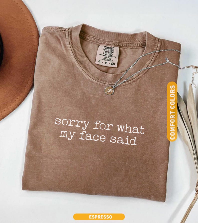 Pu&ograve; includere: T-shirt Comfort Colors color espresso con la scritta bianca "sorry for what my face said". Una collana d'argento con un ciondolo &egrave; appoggiata sulla maglietta. Un cappello marrone &egrave; visibile sullo sfondo.