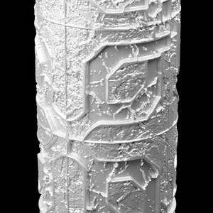 May include: A white, cylindrical object with a textured surface. The design features repeating geometric patterns and a rough, aged appearance. The background is black.