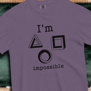 Peut inclure: T-shirt violet avec le texte "I'm impossible" et trois formes géométriques : un triangle, un carré et un cercle.