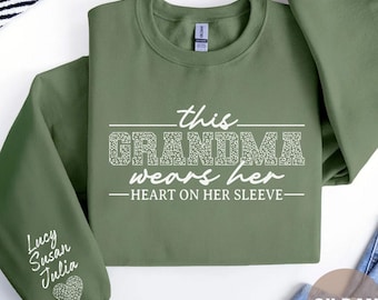 May include: Military green sweatshirt with the text "this GRANDMA wears her HEART ON HER SLEEVE" in white. The left sleeve has the names "Lucy Susan Julia" and a heart. The Gildan brand is visible.