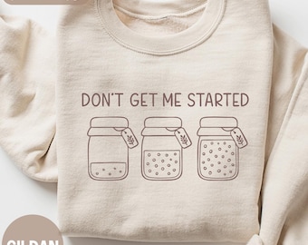 May include: Sand-colored sweatshirt with the phrase "DON'T GET ME STARTED" above a graphic of three jars. The jars have varying contents and tags. The sweatshirt is made of a soft material and is a neutral color.