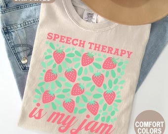 May include: Ivory-colored t-shirt with the text "SPEECH THERAPY is my jam" in pink. The shirt features a pattern of pink strawberries and green leaves. A tan hat and blue jeans are also in the image.