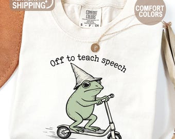 May include: Ivory-colored t-shirt featuring a cartoon frog riding a scooter, wearing a wizard hat. The text "Off to teach speech" is above the frog. The shirt has a Comfort Colors tag.