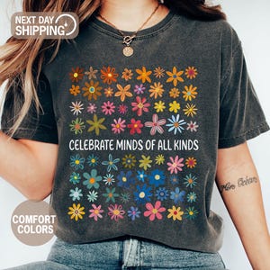 May include: Dark gray t-shirt with a colorful floral design and the text "CELEBRATE MINDS OF ALL KINDS." The shirt features a variety of flowers in shades of orange, yellow, pink, blue, and green. The shirt is made of a soft, comfortable material.