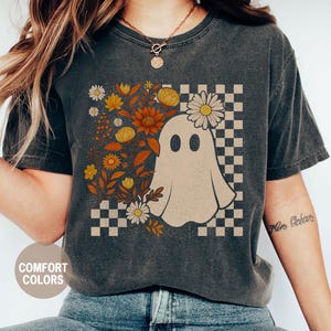 May include: Dark grey t-shirt featuring a ghost illustration with a floral design and a checkered background. The shirt has the words "Comfort Colors" and "Pepper" printed on it.