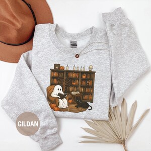 May include: Ash-colored sweatshirt featuring a Halloween-themed graphic. The design shows a ghost reading in a cozy armchair in front of a bookshelf, with pumpkins and a black cat. The sweatshirt has long sleeves and a crew neck.