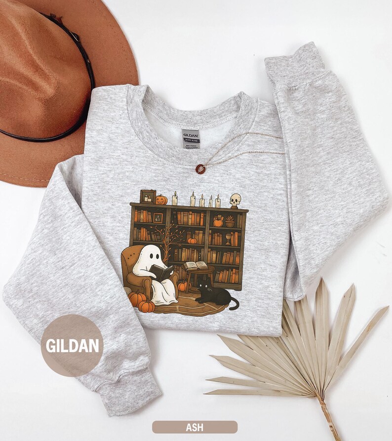 May include: Ash-colored sweatshirt featuring a Halloween-themed graphic. The design shows a ghost reading in a cozy armchair in front of a bookshelf, with pumpkins and a black cat. The sweatshirt has long sleeves and a crew neck.