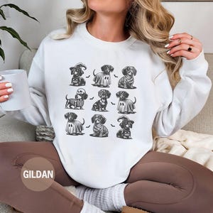 May include: White sweatshirt featuring a grid of dachshund illustrations in Halloween costumes. The dachshunds are depicted as a witch, ghost, skeleton, and more. The sweatshirt has a crew neck and long sleeves. The brand name "GILDAN" is visible.